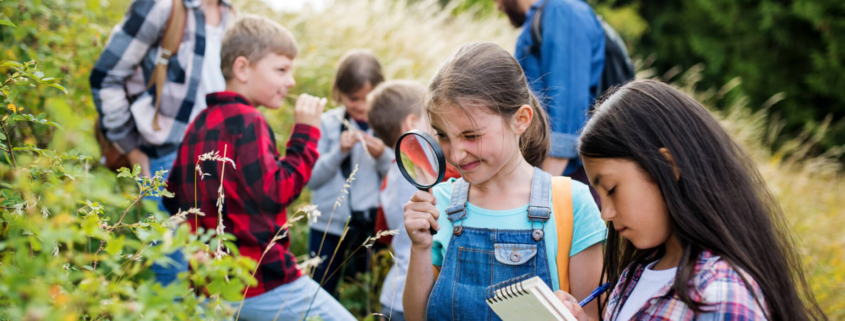 Elementary school class explores outside in a forested area. The focus is on a young girl in a blue shirt and overalls who holds a magnifying glass up to her eye in front of a plant.