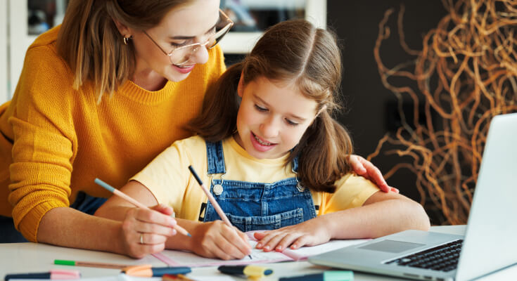 A mother in an orange sweater and glasses leans over her elementary age daughter who is working on an executive function skills practice sheet in front of her. A laptop sits open nearby.