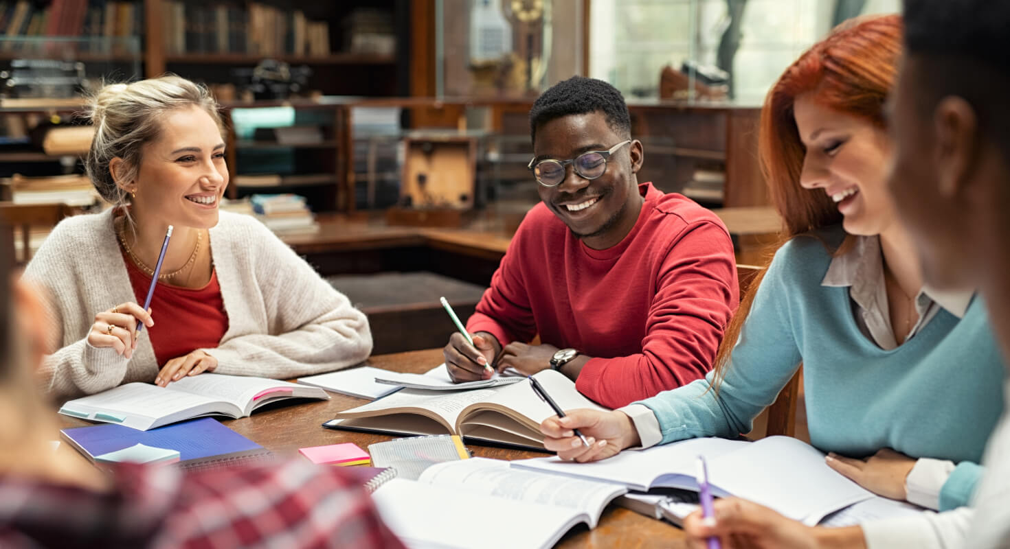 A group of young adults sit around a desk. They have schoolbooks and notebooks open in front of them and pencils in hand. They are smiling and look to be conversing with one another.