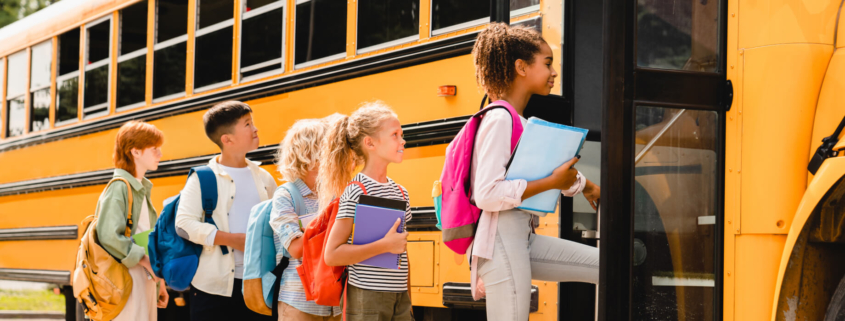 A group of elementary-aged students are standing in line waiting to board a yellow school bus. They are wearing backpacks, holding binders and folders, and look excited.