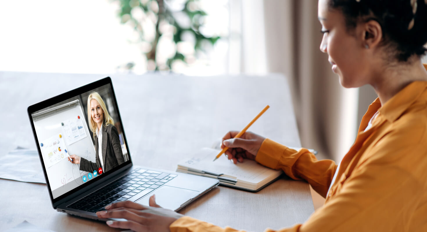 A female high school student sits at a table at home with her laptop open and a notebook to take notes. The laptop plays a video of a teacher providing academic support.