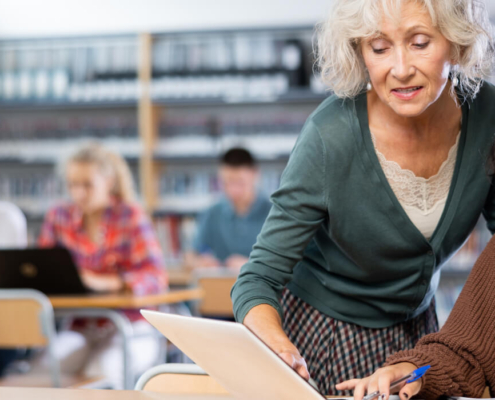 A female educator with white hair and a green sweater leans over a high school student's desk, pointing at a laptop and providing academic support.