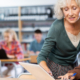 A female educator with white hair and a green sweater leans over a high school student's desk, pointing at a laptop and providing academic support.