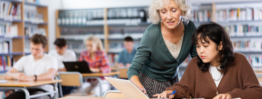 A female educator with white hair and a green sweater leans over a high school student's desk, pointing at a laptop and providing academic support.