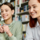 Middle school female student sits next to her smiling academic coach in a library setting as they look at her work together.