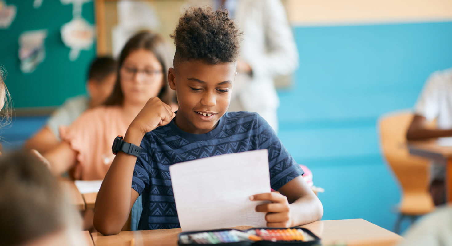 A middle school aged, Black male student sits in class looking at a piece of paper he holds. He looks happy, succeeding after he received specialized academic support.