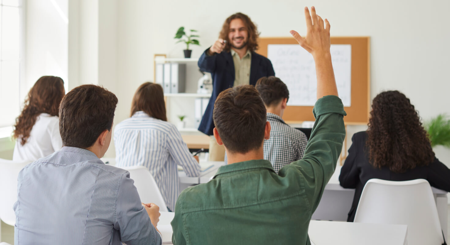 A group of high school students sit in a classroom with a young male teacher standing at the front. The teacher smiles and calls on a boy who is raising his hand.