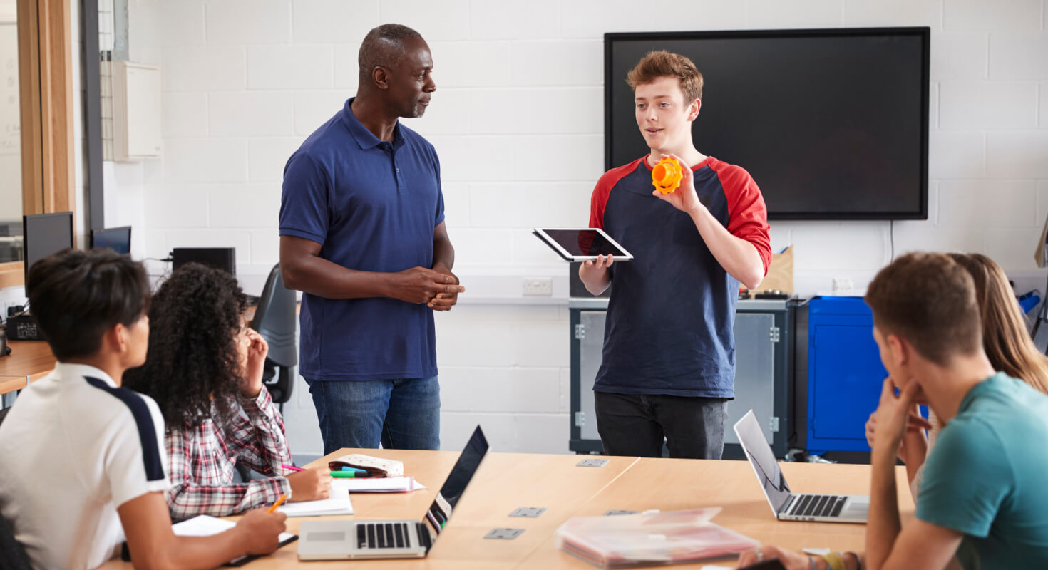 High school students gather, with one student and an educator standing as they talk and study together.