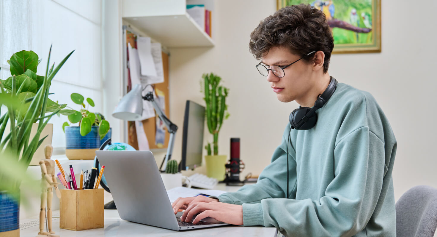 Male high school student in light blue sweatshirt sits at home in a well-organized study environment, focused on his work.