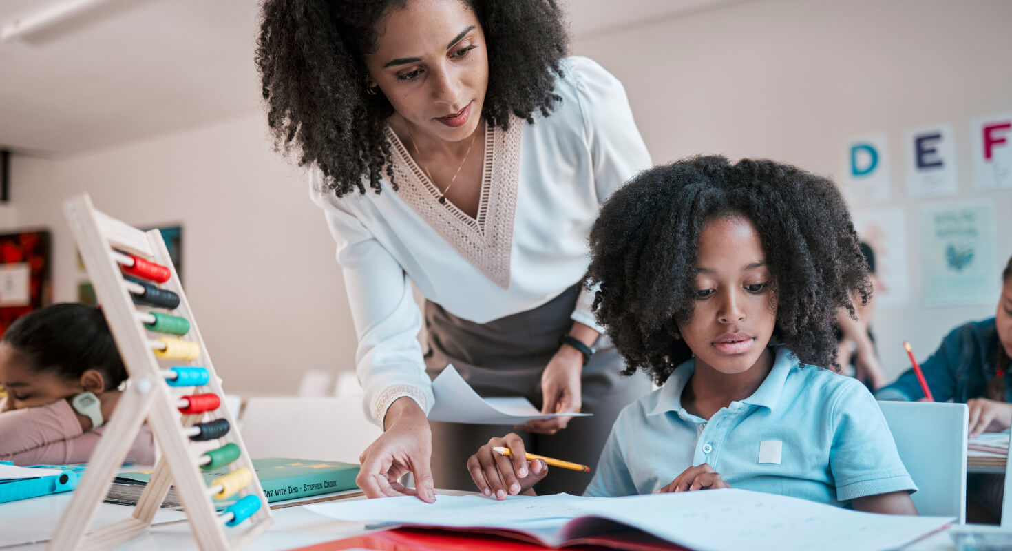 A young Black female teacher assists a Black elementary school student in the classroom in an effort to create a structured environment.