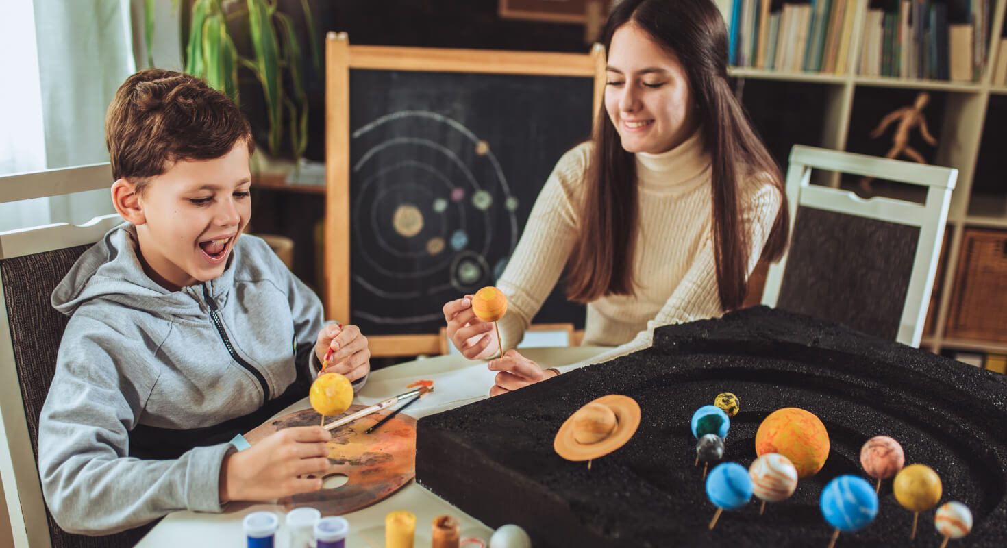 An educator sits with an elementary school student as they work on a hands-on science project about the solar system.