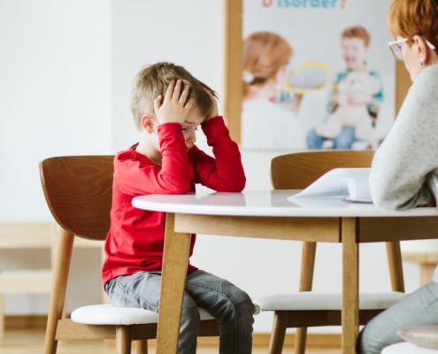 Young male student in a red shirt sits at a classroom table with an educator. The student is frustrated and resting his head in his hands.