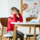 Young male student in a red shirt sits at a classroom table with an educator. The student is frustrated and resting his head in his hands.