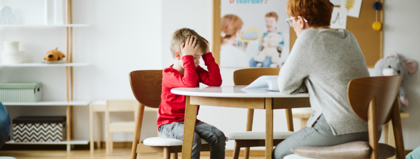 Young male student in a red shirt sits at a classroom table with an educator. The student is frustrated and resting his head in his hands.