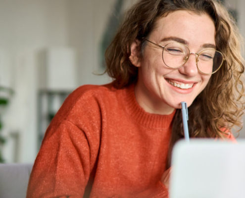 High school female student sits smiling at her laptop at home, holding a pen to her chin as she practices good study habits