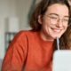 High school female student sits smiling at her laptop at home, holding a pen to her chin as she practices good study habits