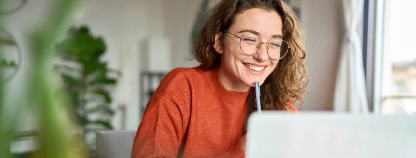 High school female student sits smiling at her laptop at home, holding a pen to her chin as she practices good study habits