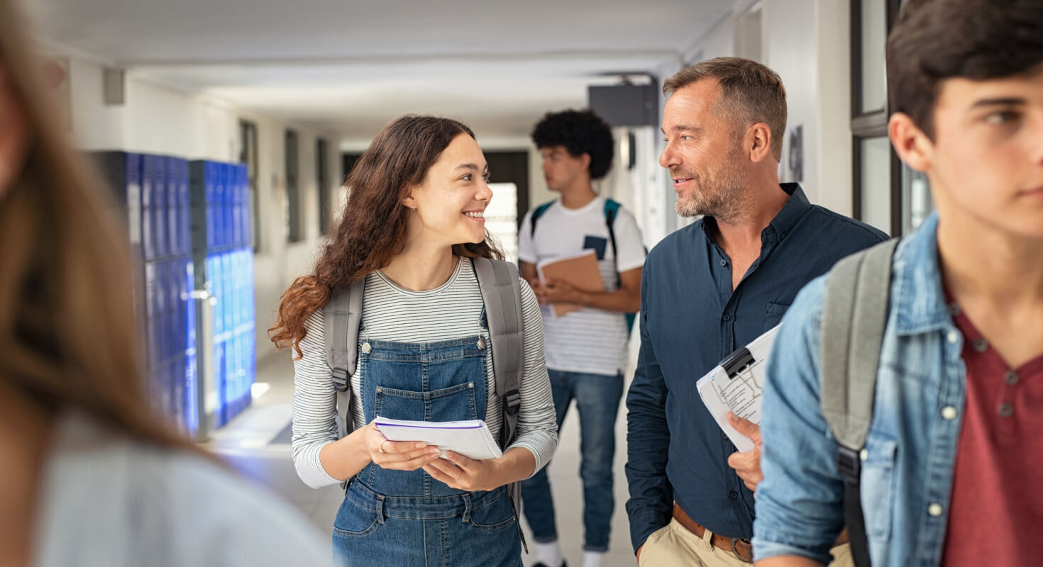 A high school female student smiles and walks down a school hallway talking to her academic coach who is holding a stack of papers. Other students walk around them.