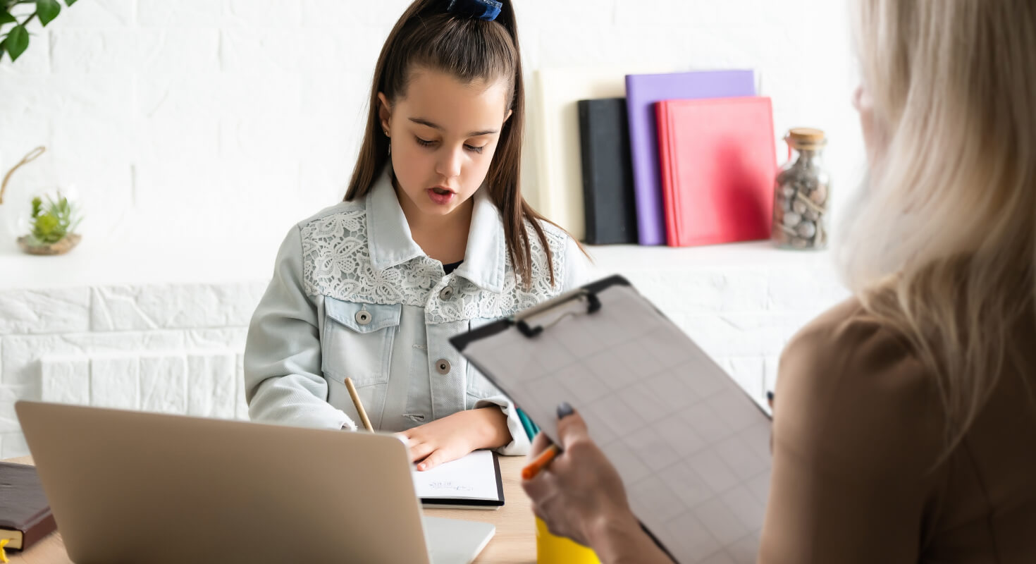 Elementary age girl sits at a table with an ADHD coach in the foreground. She works on writing her goals as a learning technique.