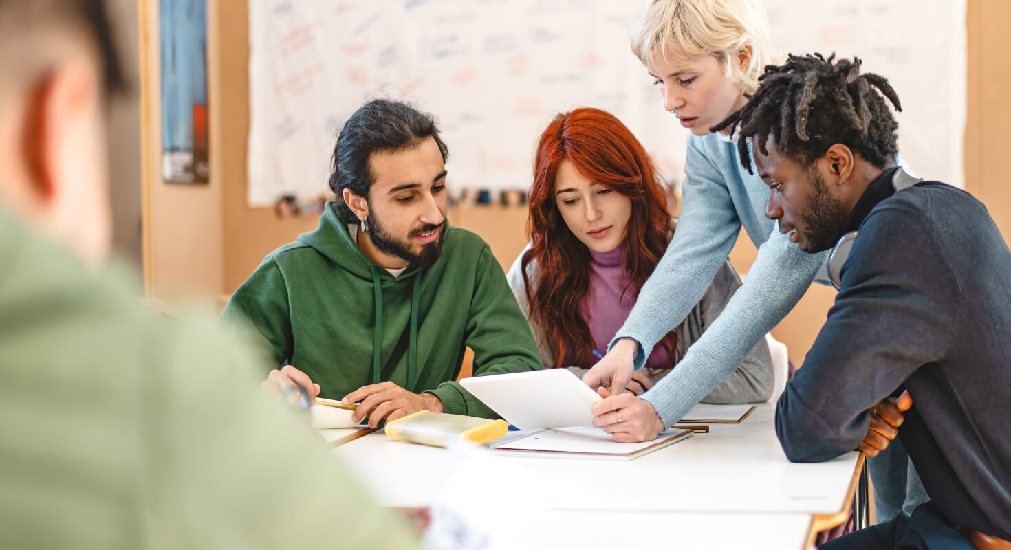 Study group of three people sit at a table with an academic coach leaning across the table to point at a key study point on a piece of paper.