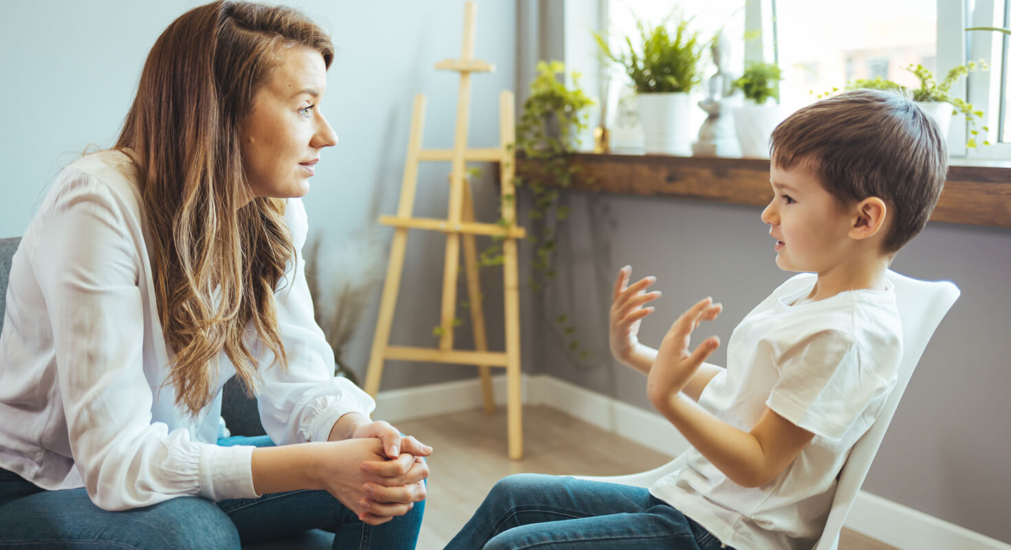 A female ADHD coach sits with a young elementary school age boy as he gestures and talks, explaining his behavior to her.