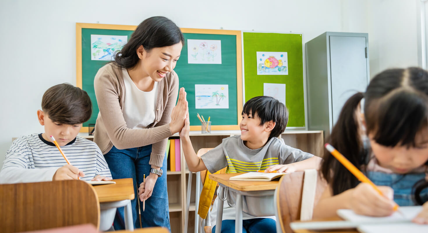 A female teacher high fives an elementary school student as he sits in his seat, showing an example of positive reinforcement in the classroom.