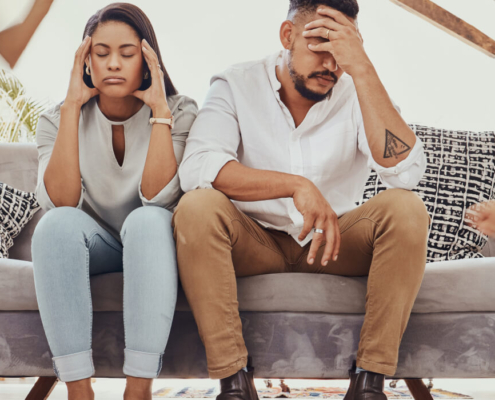 Parents sit on the couch with their heads in their hands looking frustrated while children run around them