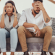 Parents sit on the couch with their heads in their hands looking frustrated while children run around them