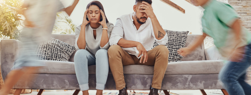 Parents sit on the couch with their heads in their hands looking frustrated while children run around them