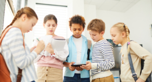 A group of young children at school gather around a young boy holding a cellphone.