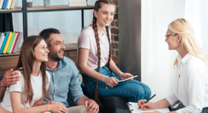 Two parents sit close to one another while they and their daughter smile at a woman writing notes on a clipboard.