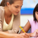 An adult helps a young child in a pink shirt with a written assignment