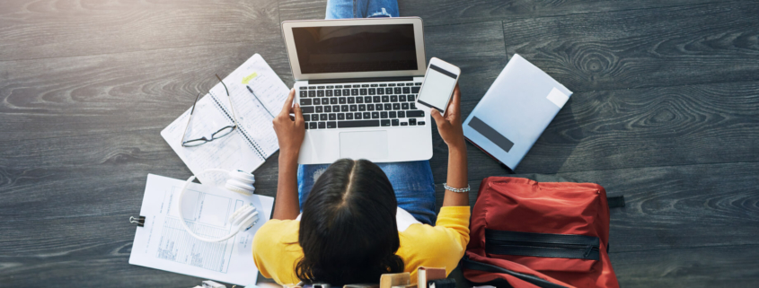 A student sits on the floor among a laptop and paperwork.